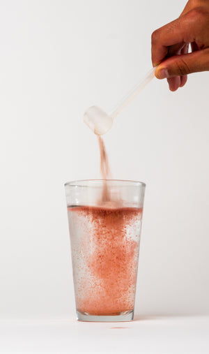 A hand holding a clear scoop of iAMINOS Hydrate+. pouring a fine pink powder into a clear glass of water, creating a bubbly orange liquid, on a clean white background.