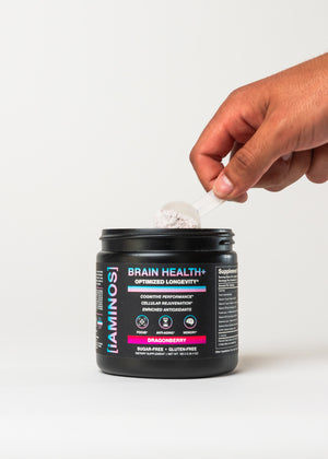 A hand holding a white scoop above a black tub of iAMINOS Brain Health+ dietary supplement powder on a clean white background.