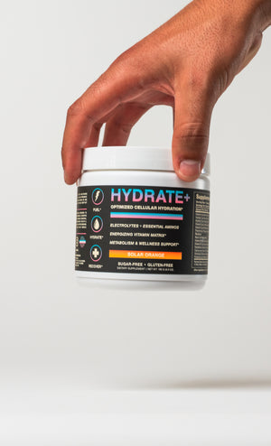 A hand holding a white tub of iAMINOS Hydrate+ electrolyte supplement powder against a clean white background.