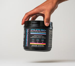 A hand holding a black tub of iAMINOS Athlete Pro+ performance powder against a clean light gray background.