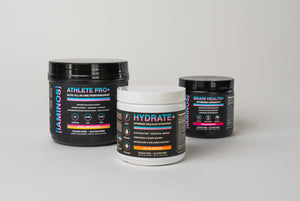 Three supplement containers from iAMINOS on a light gray studio background: a large black Athlete Pro+ tub, a smaller black Brain Health+ tub, and a white Hydrate+ tub in the foreground.