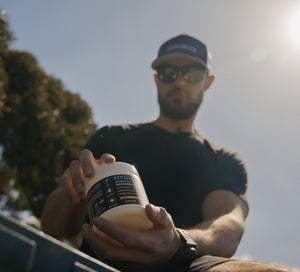 A man outdoors wearing a black cap and sunglasses, holding a white tub of iAMINOS Hydrate+ electrolyte and wellness supplement. The background features bright sunlight and green foliage.