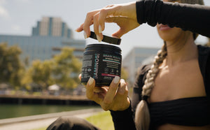 A woman outdoors holds an open black supplement container labeled iAMINOS Brain Health+. The background shows a blurred city waterfront scene with buildings and greenery.