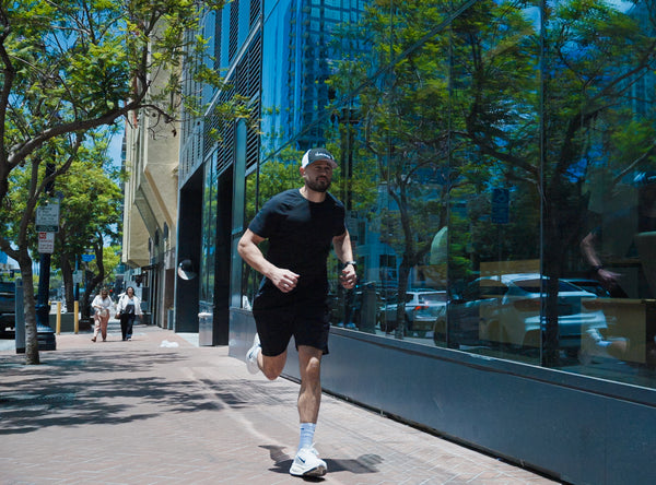 A man running on a city sidewalk next to a modern glass building, wearing an iAMINOS hat, black athletic t-shirt, black shorts, white socks, and white running shoes.