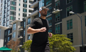 A man jogging outdoors in a black athletic t-shirt and black shorts, wearing a black and white trucker hat with a visible iAMINOS logo. The background shows modern high-rise apartment buildings and green trees under bright daylight.