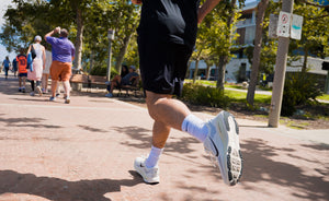 A runner's legs in black athletic shorts, white socks, and white running shoes jogging on a paved path in a sunny park with trees and other joggers in the background.